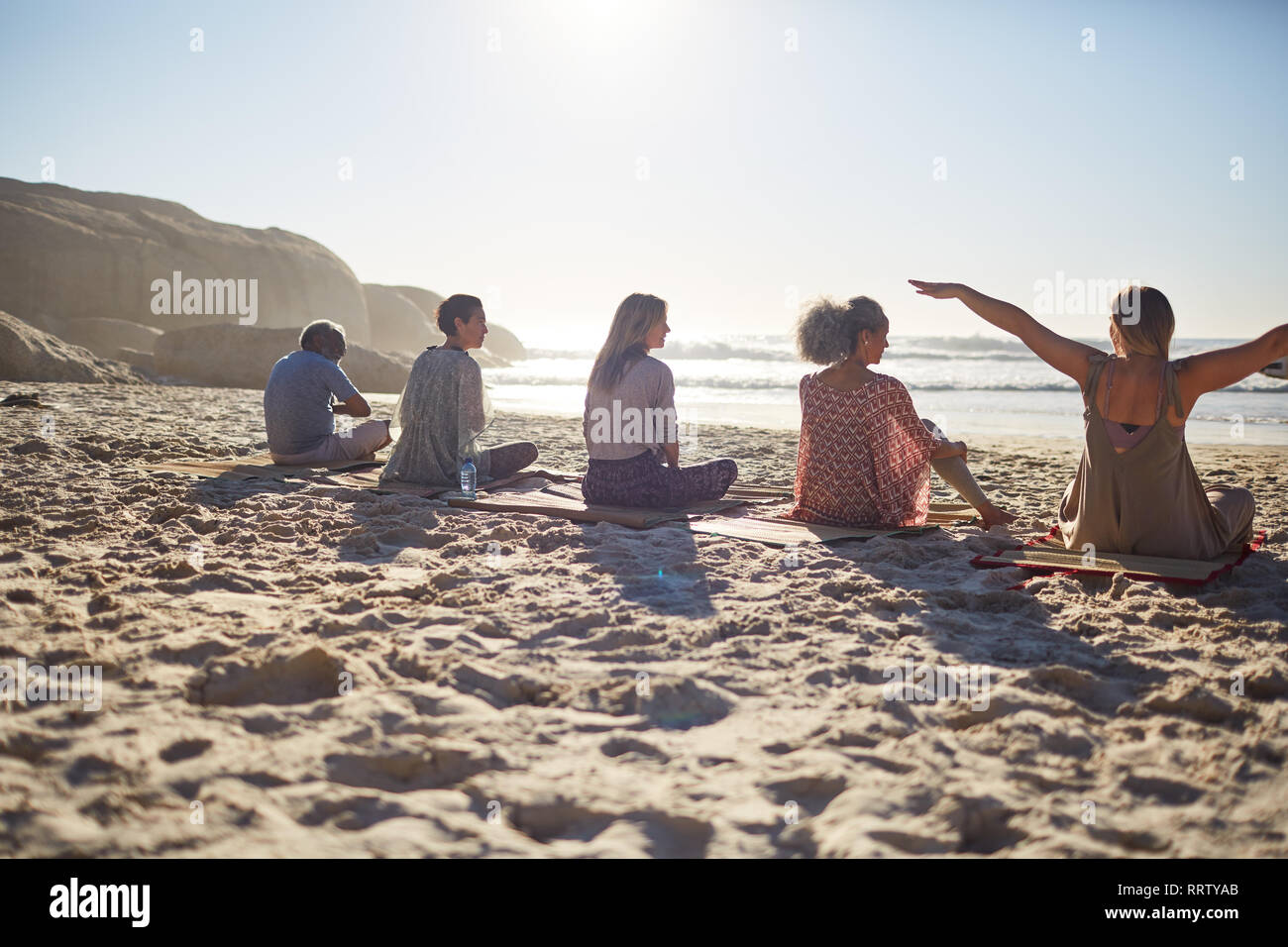 Group sitting on yoga mats on sunny beach during yoga retreat Stock Photo