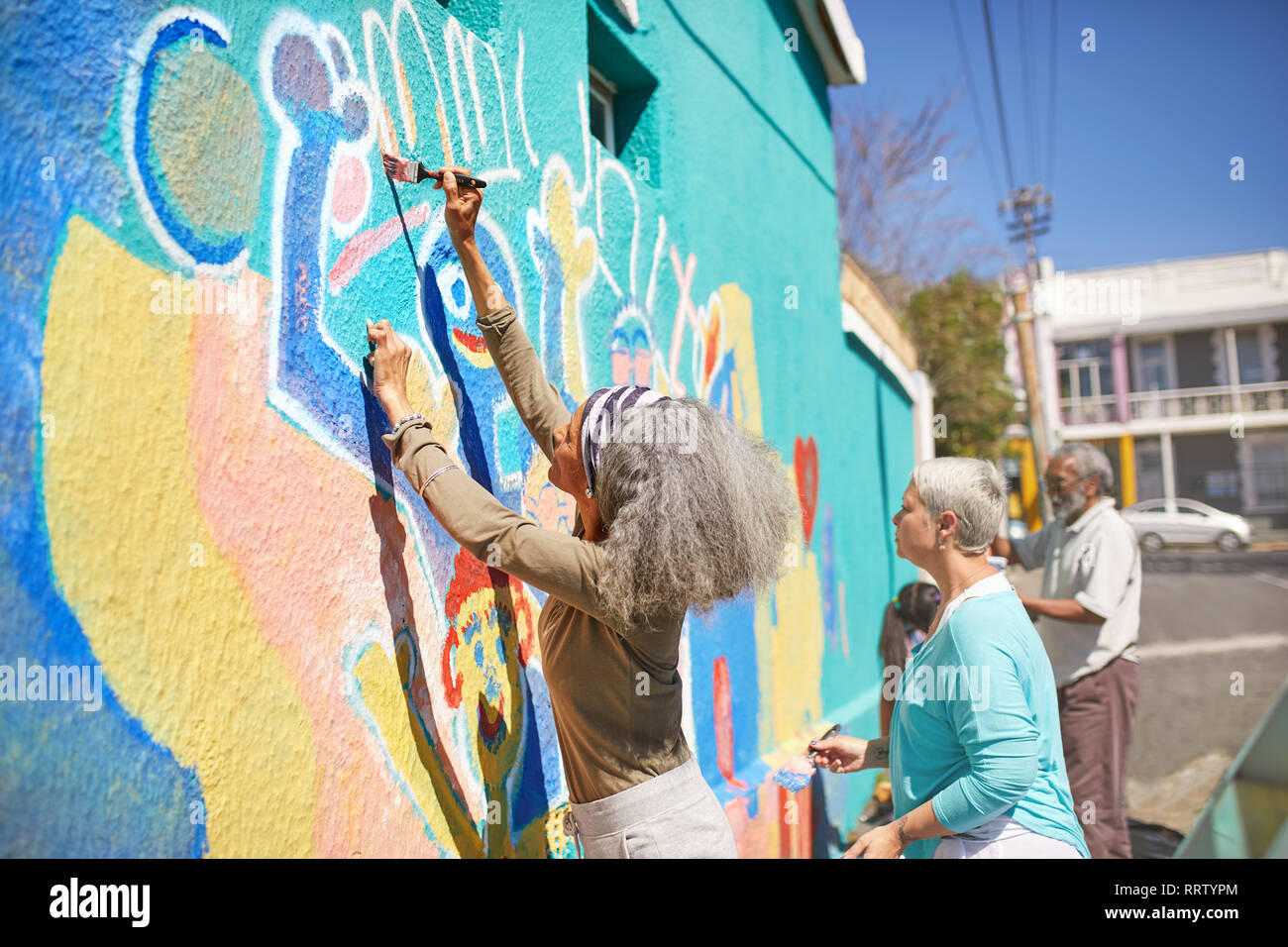 Senior community volunteers painting vibrant mural on sunny urban wall Stock Photo