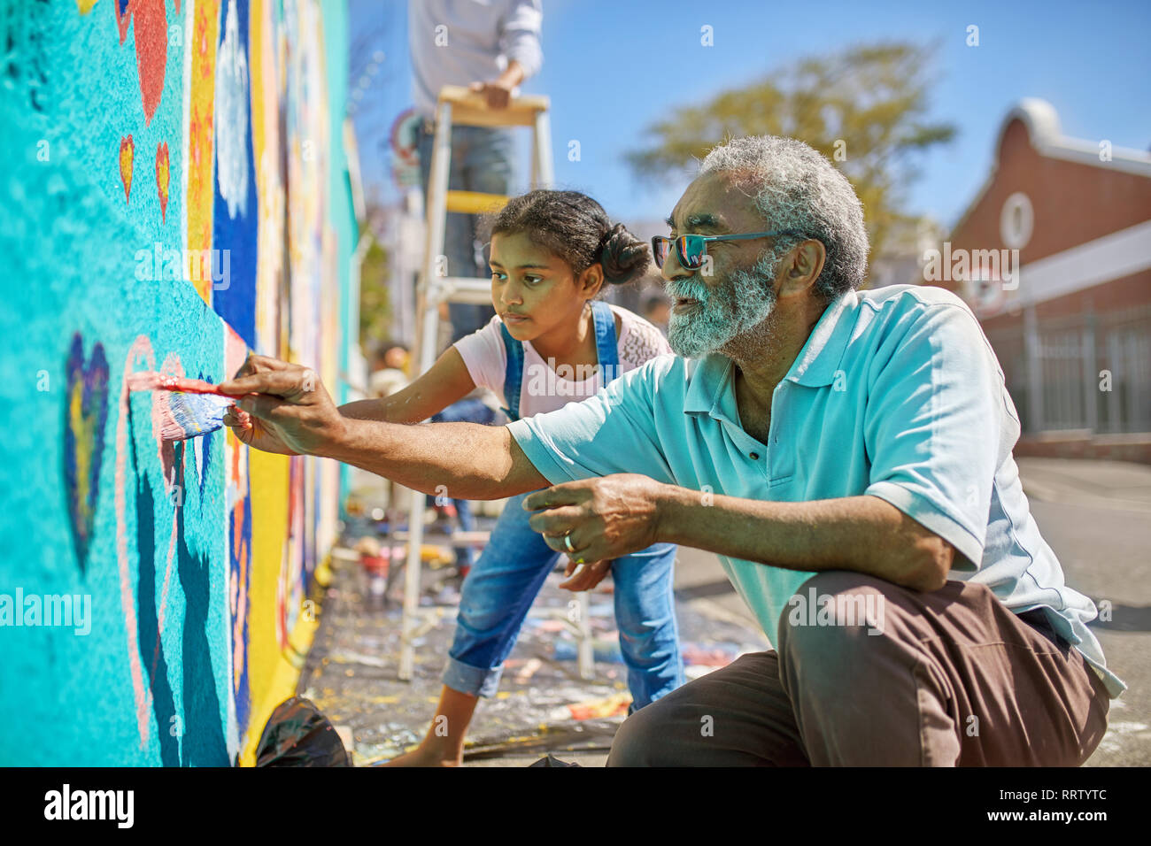 Grandfather and granddaughter volunteers painting vibrant mural on sunny urban wall Stock Photo
