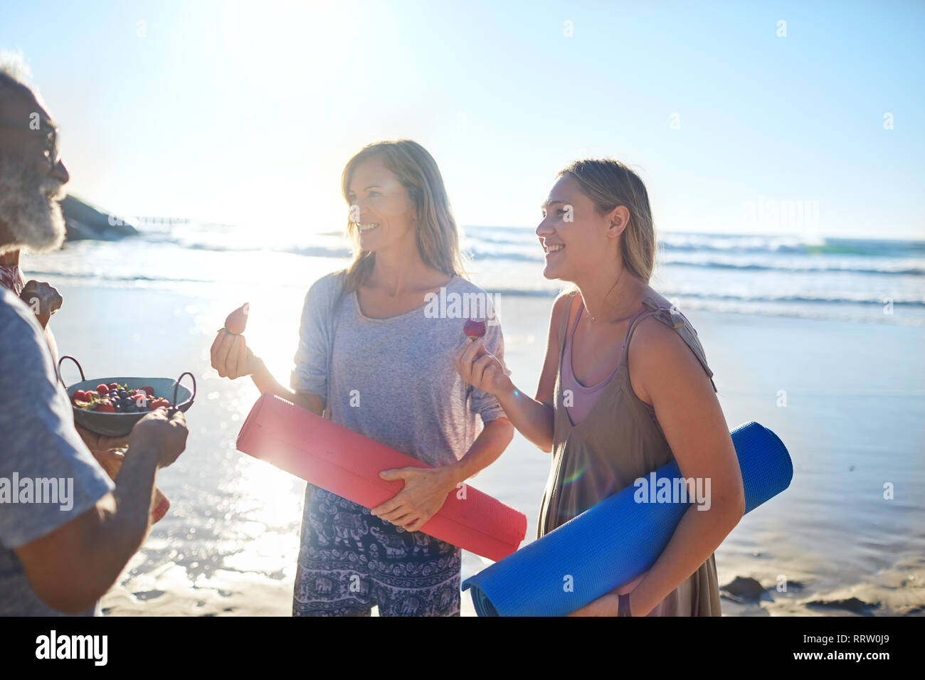 Mother and daughter with yoga mats eating fresh berries on sunny beach during yoga retreat Stock Photo