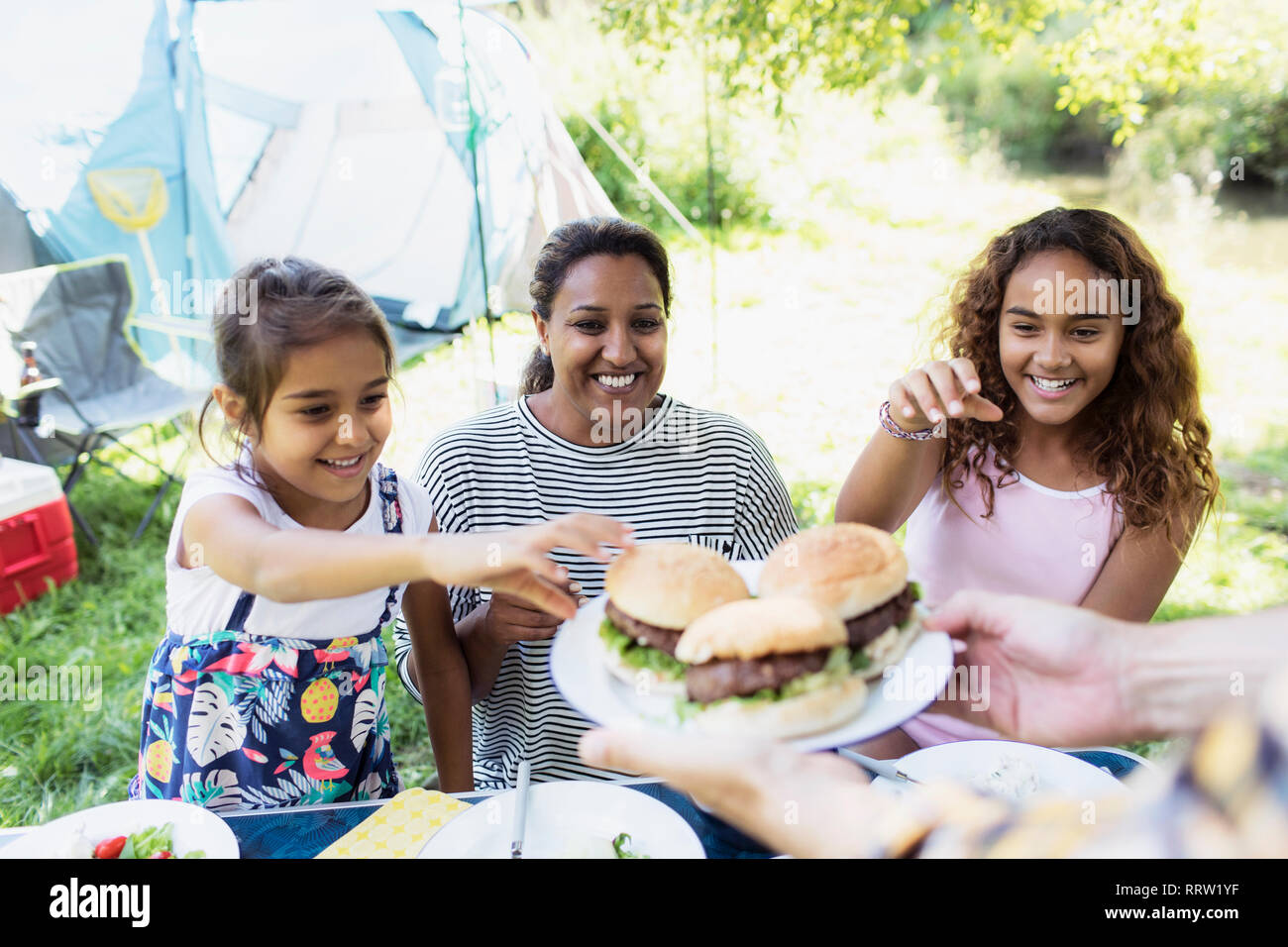 Happy mother and daughters reaching for barbecue hamburgers at campsite Stock Photo