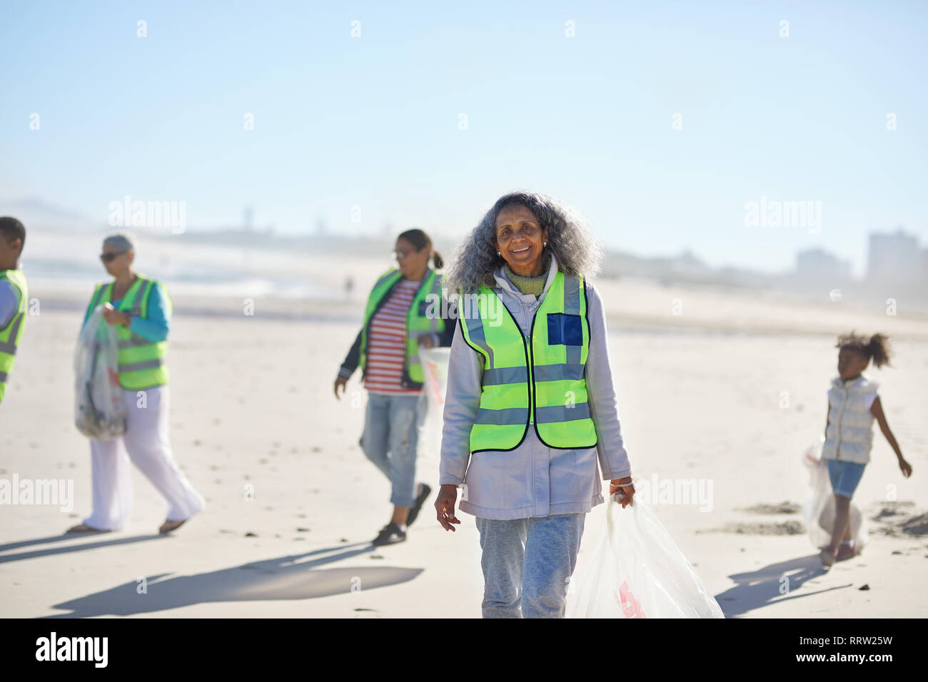 Portrait confident senior woman volunteer cleaning up litter on sunny beach Stock Photo