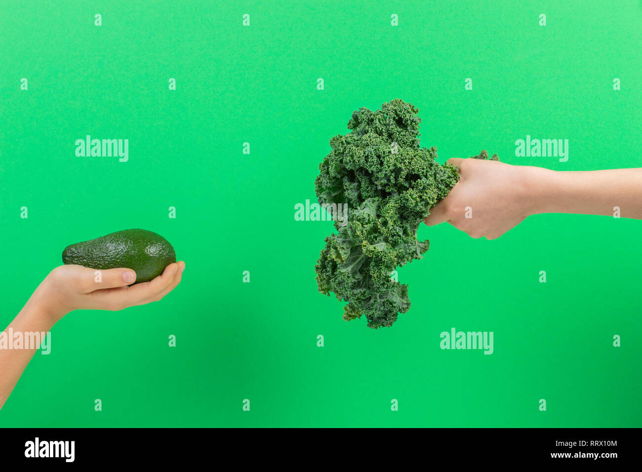 Healthy vegetables for kids. Kid hand with bunch of kale leaves and kid hand holding avocado over green background Stock Photo