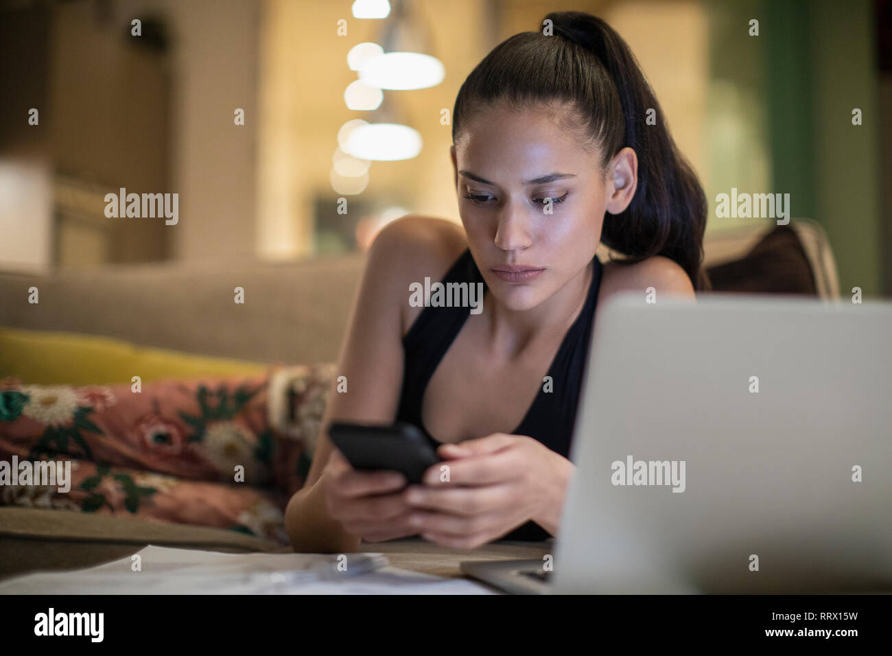 Focused woman in pajamas using smart phone at laptop on sofa Stock Photo