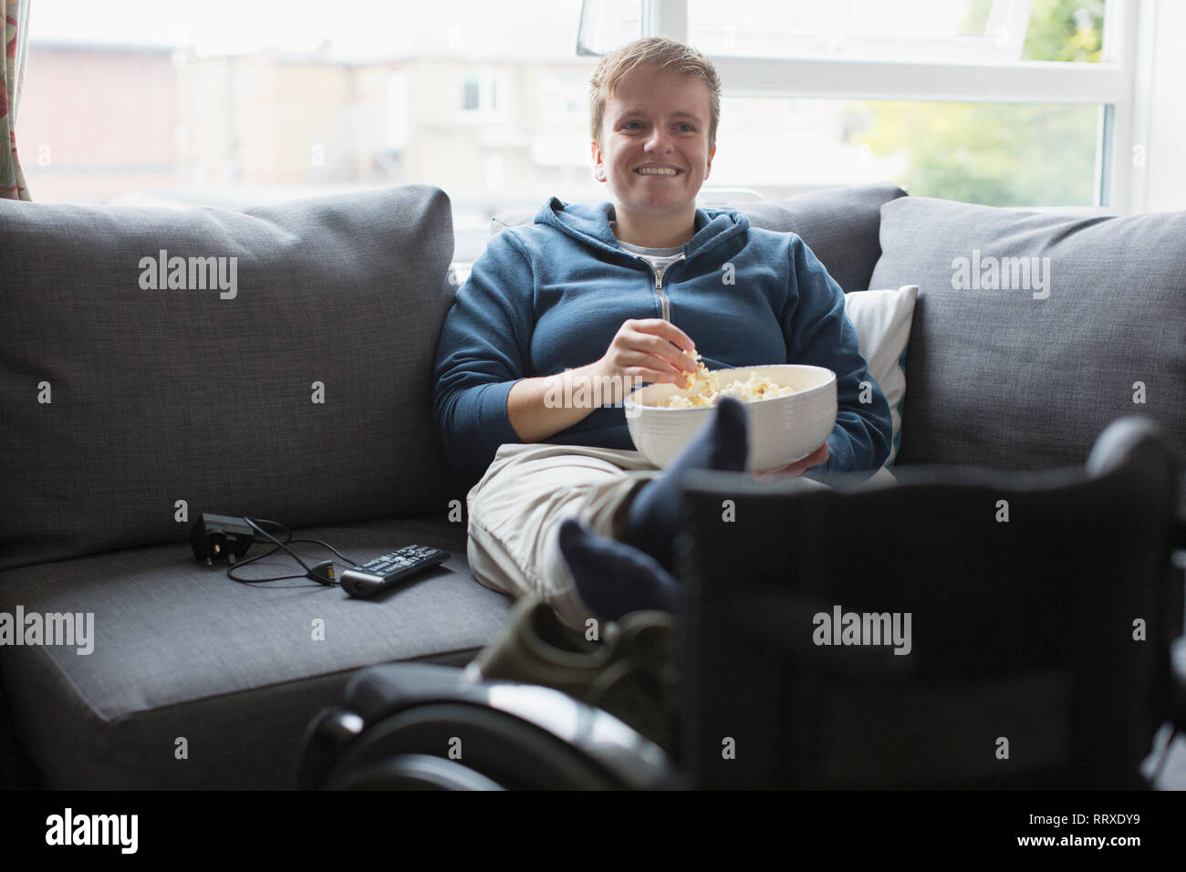 Smiling young woman watching TV and eating popcorn on sofa with feet up on wheelchair Stock Photo