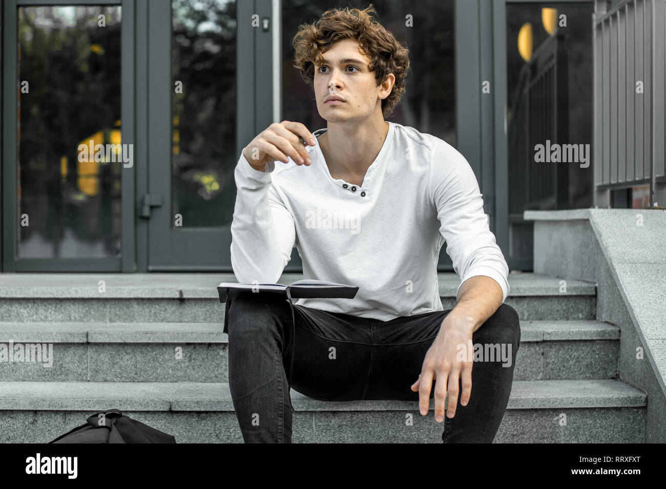 Attractive pensive student sitting on modern building steps and making notes in his black notepad. Stock Photo