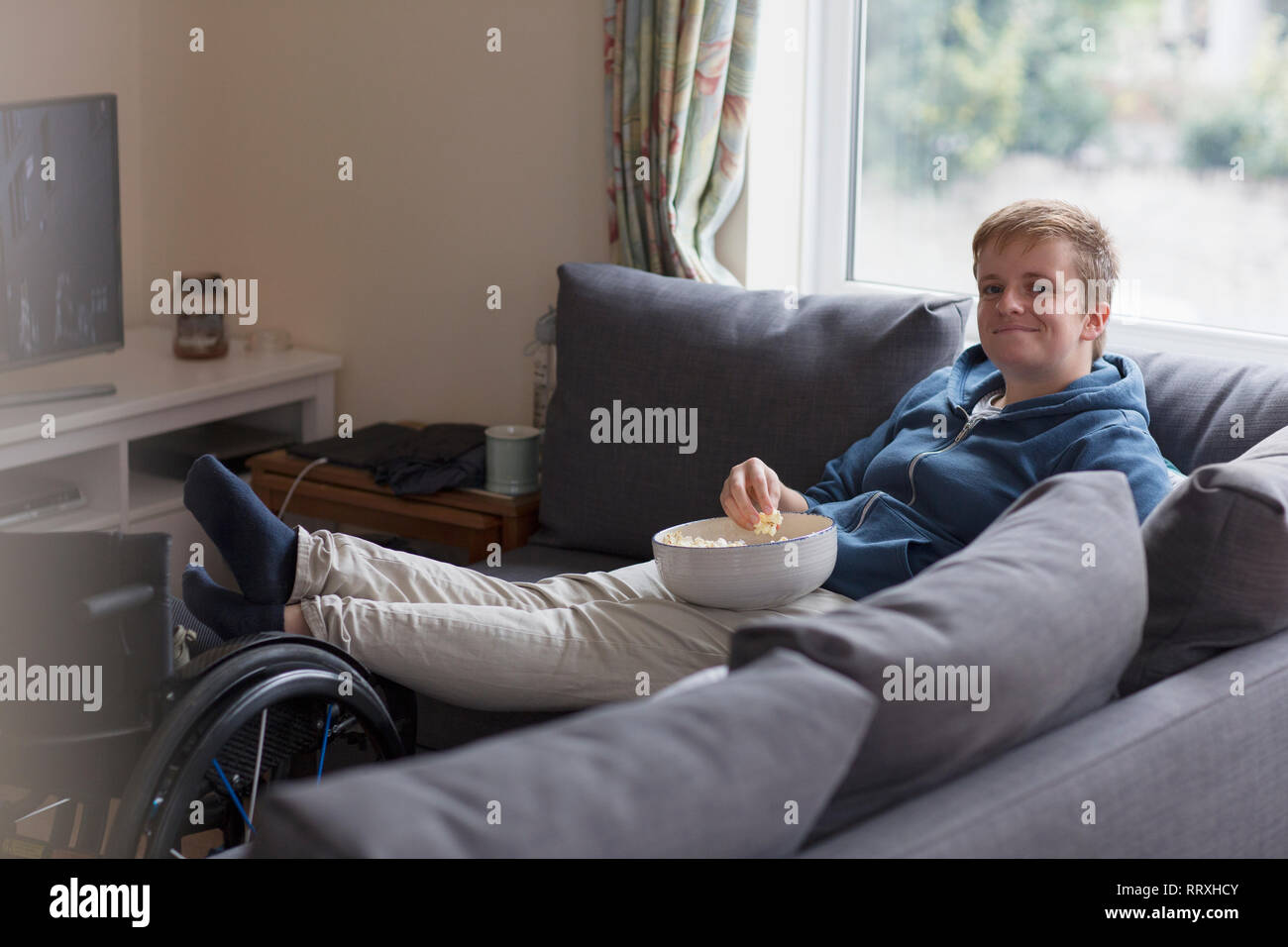 Portrait confident young woman watching TV and eating popcorn on sofa with feet up on wheelchair Stock Photo