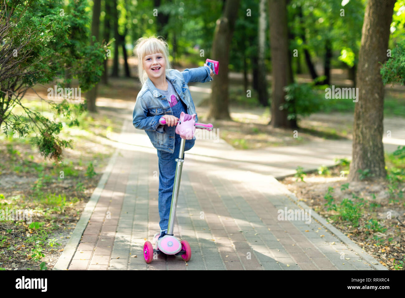 Cute little blond kid girl riding scooter in city park ...