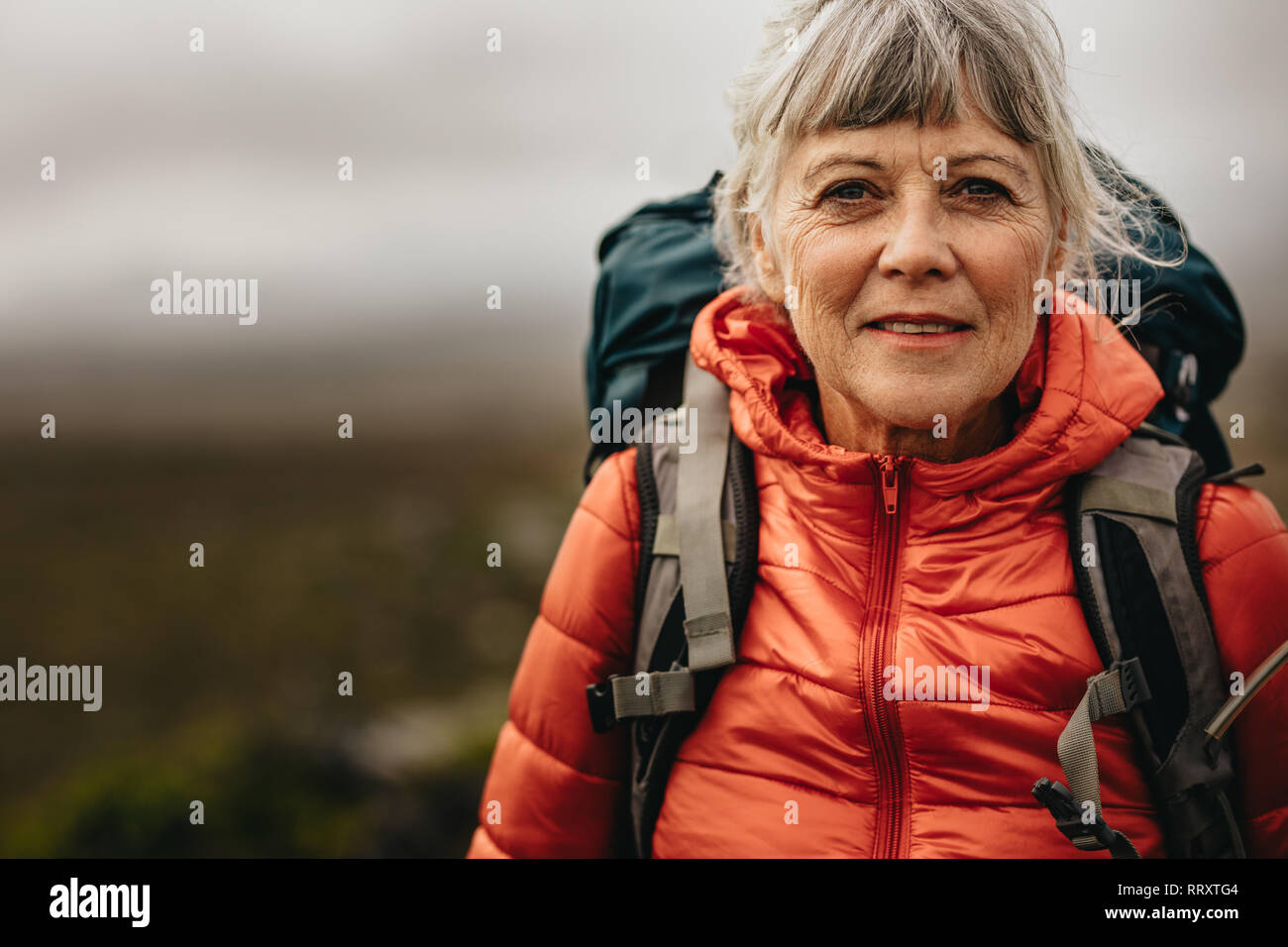 Portrait of a senior woman standing outdoors on a holiday. Senior woman wearing a backpack and jacket standing during a trekking trip. Stock Photo