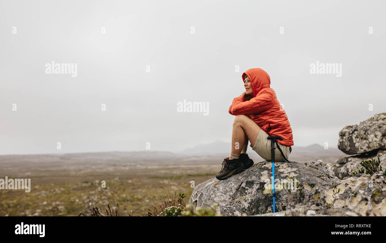Woman sitting on a rock on a hilltop looking at the beauty of nature. Senior woman sitting relaxed after her trek with a hiking pole by her side. Stock Photo