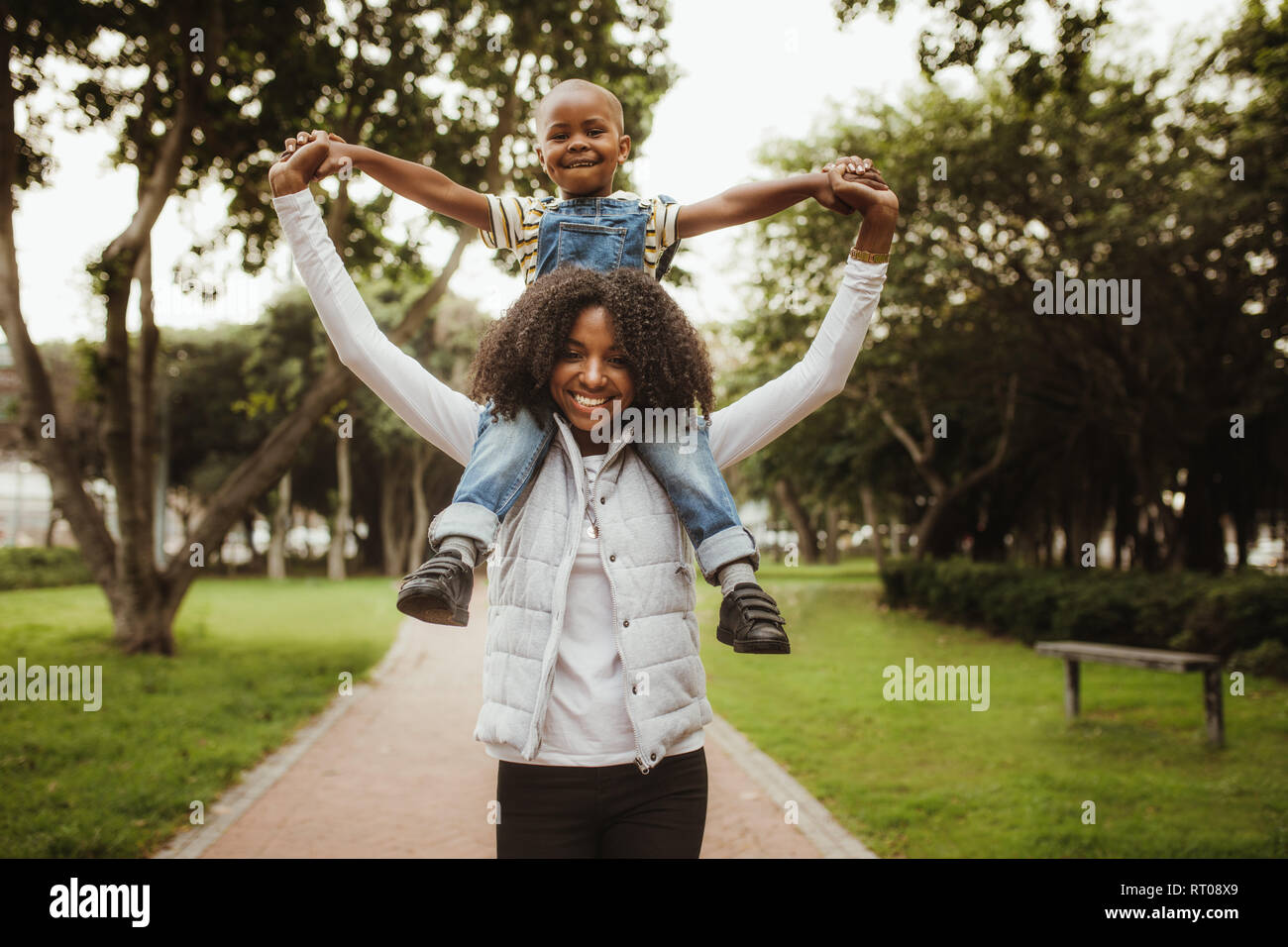Happy mother giving son a piggyback ride on her shoulders in park. African woman carrying his boy on her shoulders while walking in a park. Stock Photo
