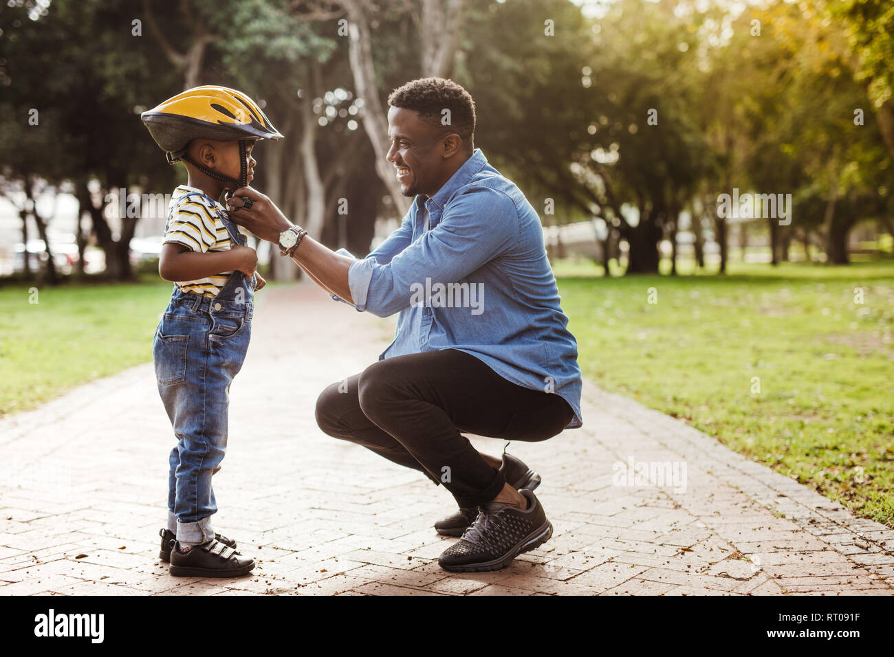 African man putting helmet on cute boy at the park. Father puts his son a protective helmet for riding bike. Stock Photo