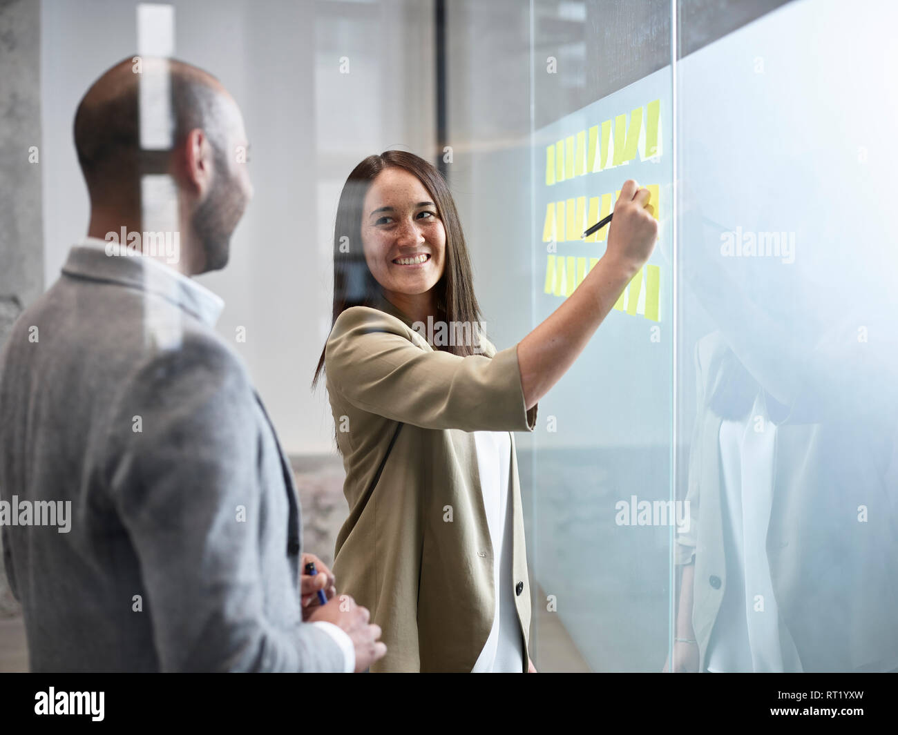 Businesswoman smiling at colleague writing on sticky notes at glass pane in office Stock Photo