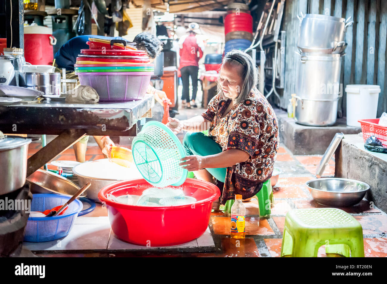 An Old Cambodian Lady Scrubs All The Colourful Dishes And Buckets