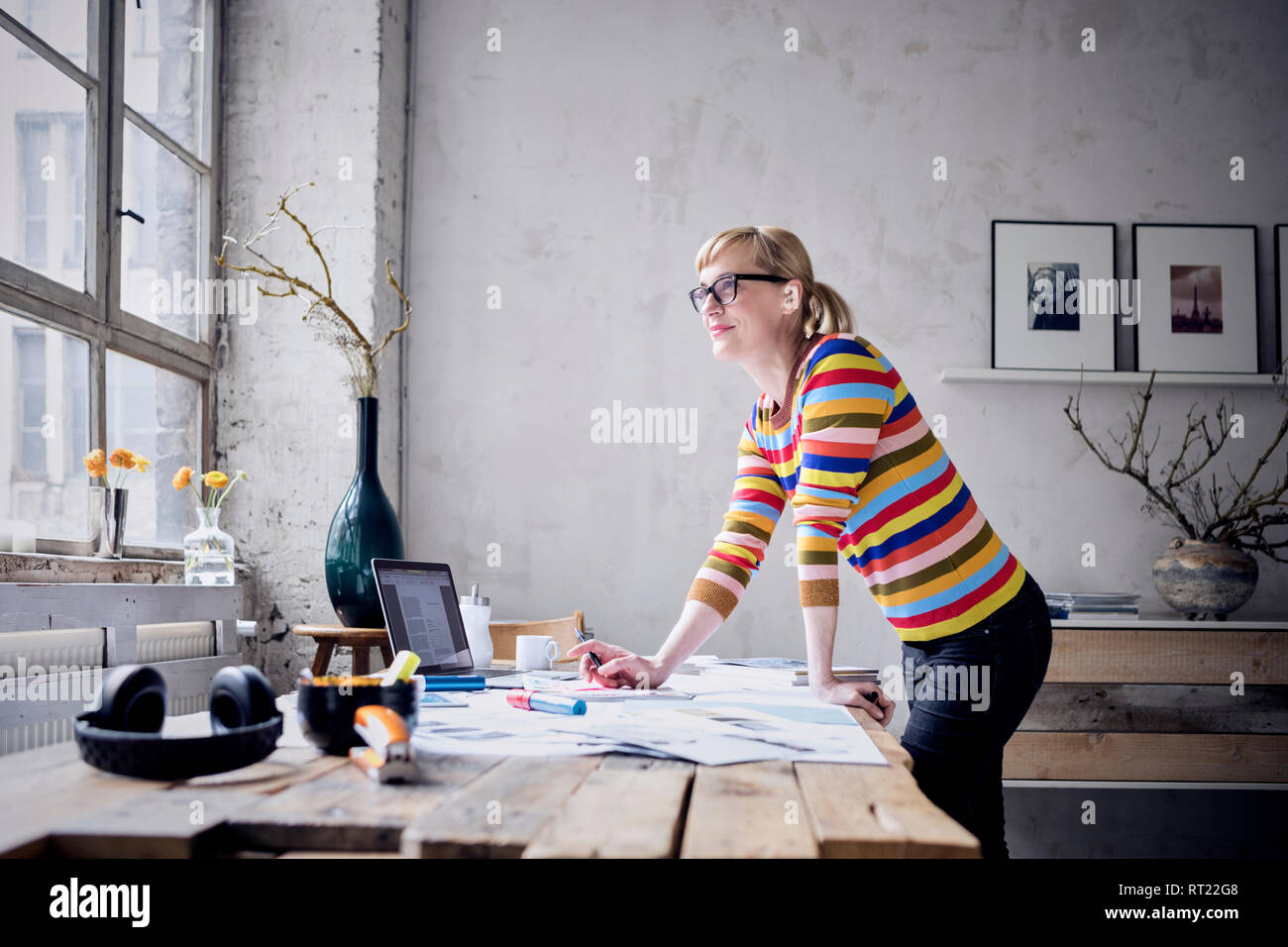 Portrait of smiling woman standing at desk in a loft looking through window Stock Photo