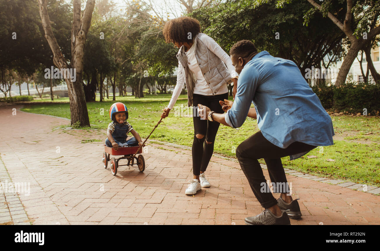 Boy sitting in a small wagon pulled by his mother, while father standing with his arms outstretched. Young family on enjoying at park. Stock Photo