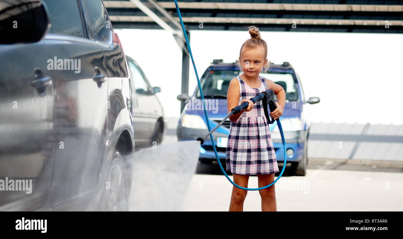 Horizontal image pretty little preschool girl in summer dress helping to parents use coin-operated self-serve car wash, clean auto concept Stock Photo