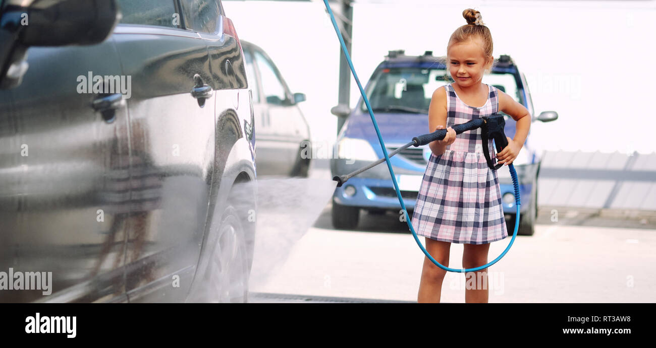 Horizontal view pretty little preschool girl in summer dress helping to parents use coin-operated self-serve car wash Stock Photo