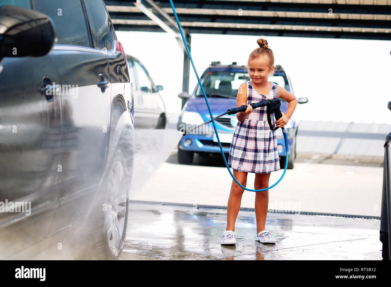 Pretty little preschool girl in summer dress helping to parents use coin-operated self-serve car wash, cleaning auto exterior Stock Photo