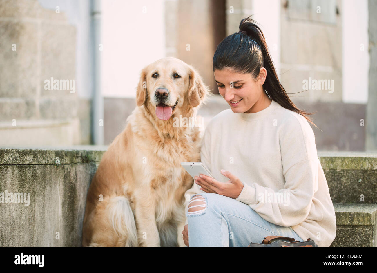 Young woman with her Golden retriever dog on stairs outdoors using cell phone Stock Photo