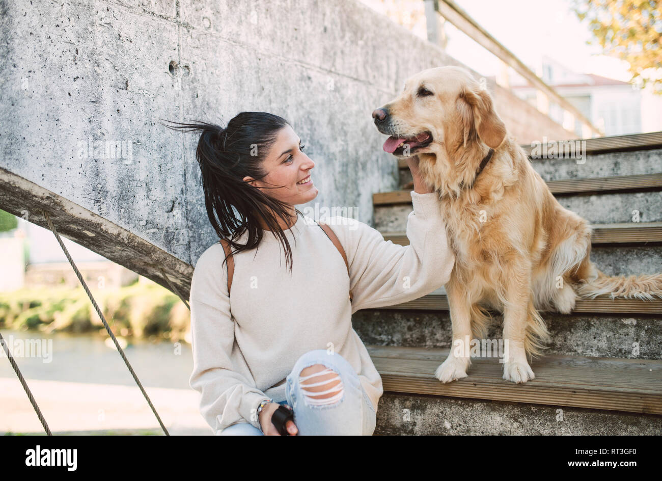 Smiling young woman stroking her Golden retriever dog on stairs outdoors Stock Photo