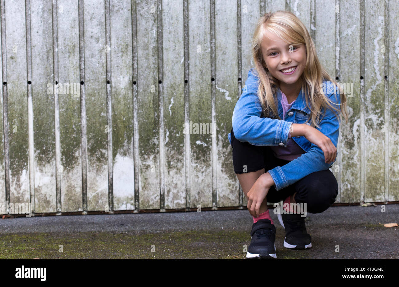 Portrait of smiling blond girl crouching in front of wooden wall Stock Photo