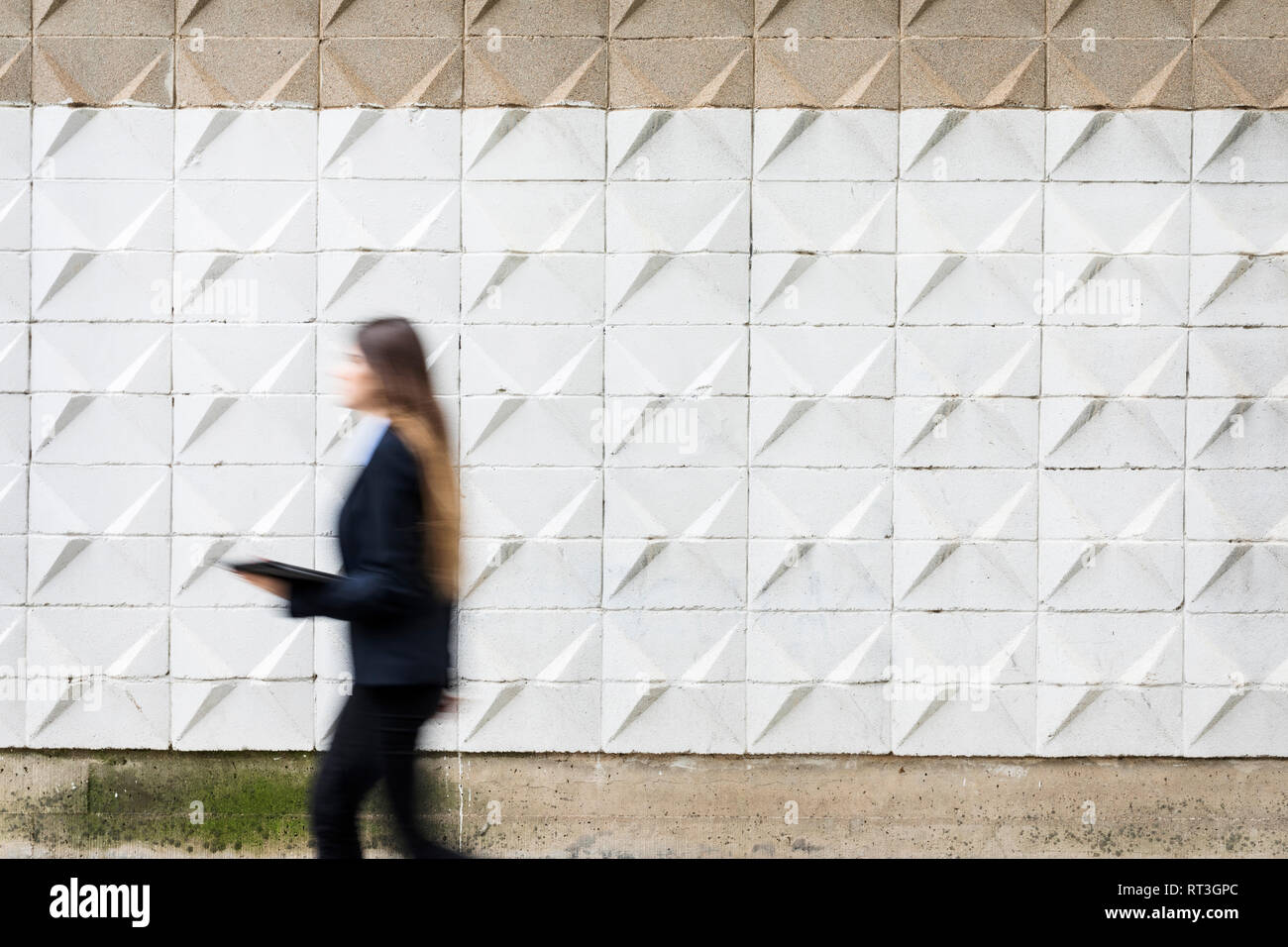 Blurred young businesswoman holding tablet passing concrete wall Stock Photo
