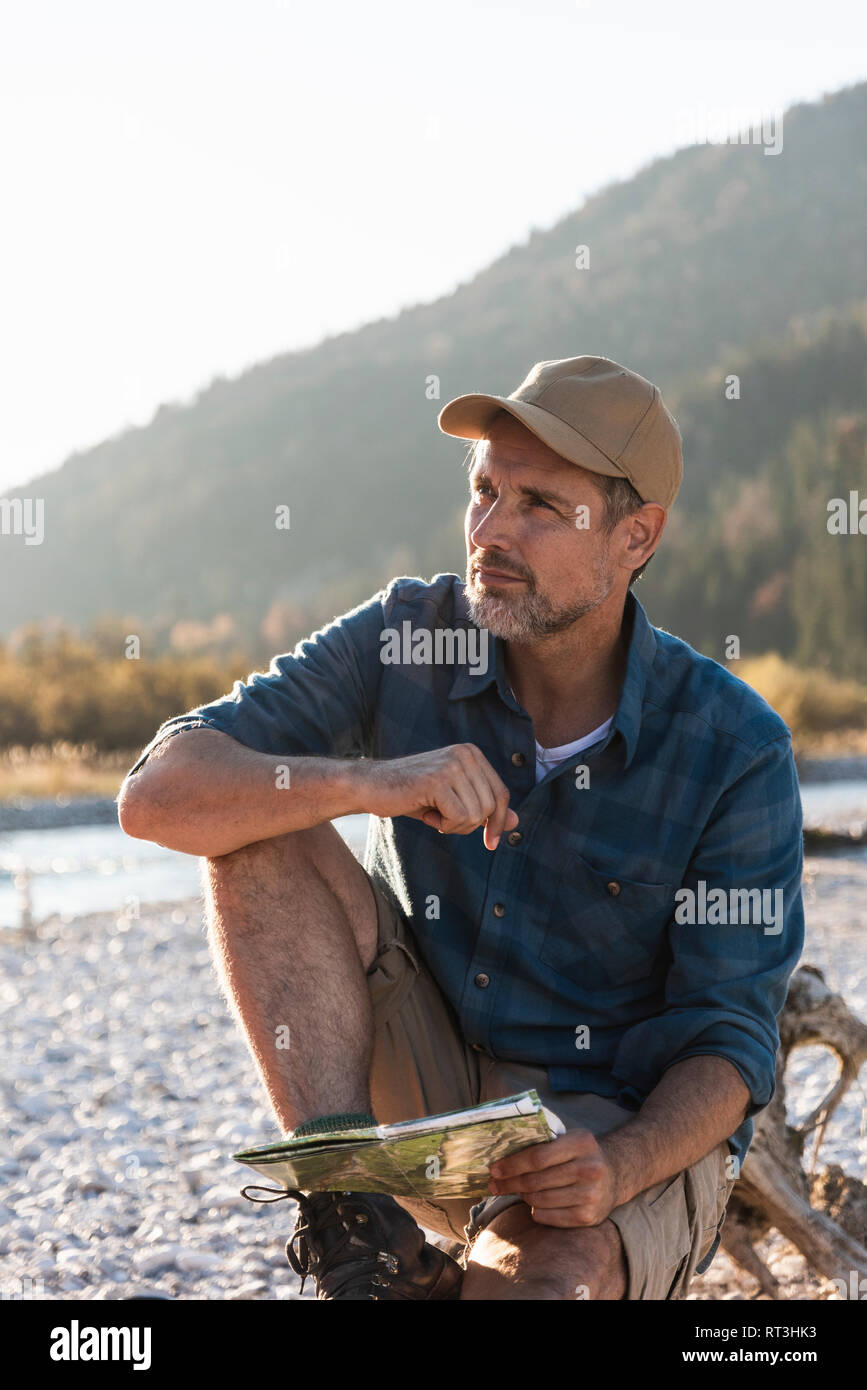 Mature man camping at riverside, sitting on tree trunk with map Stock Photo