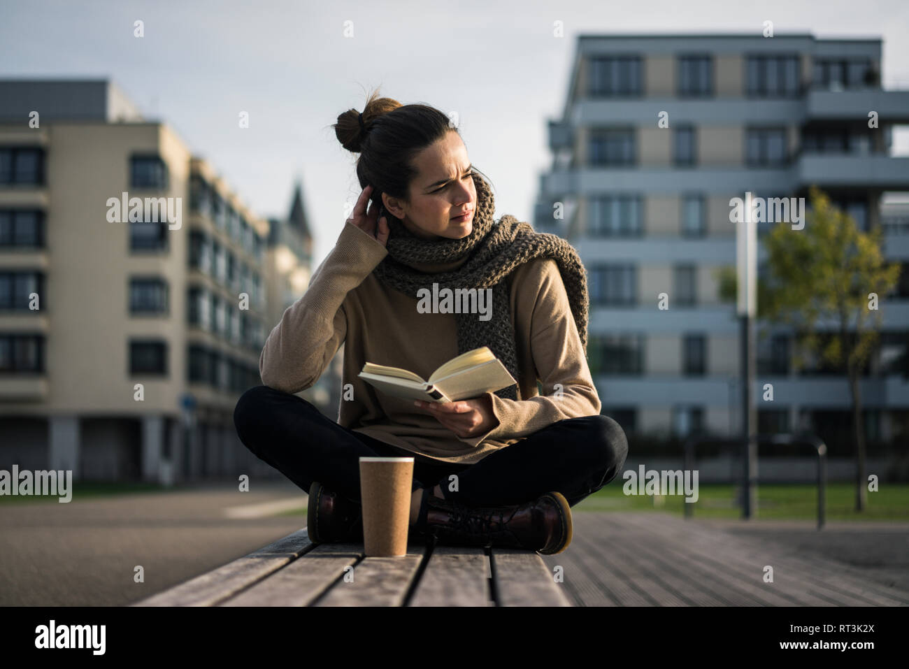 Melancholy woman with coffee to go and book sitting on bench in autumn Stock Photo