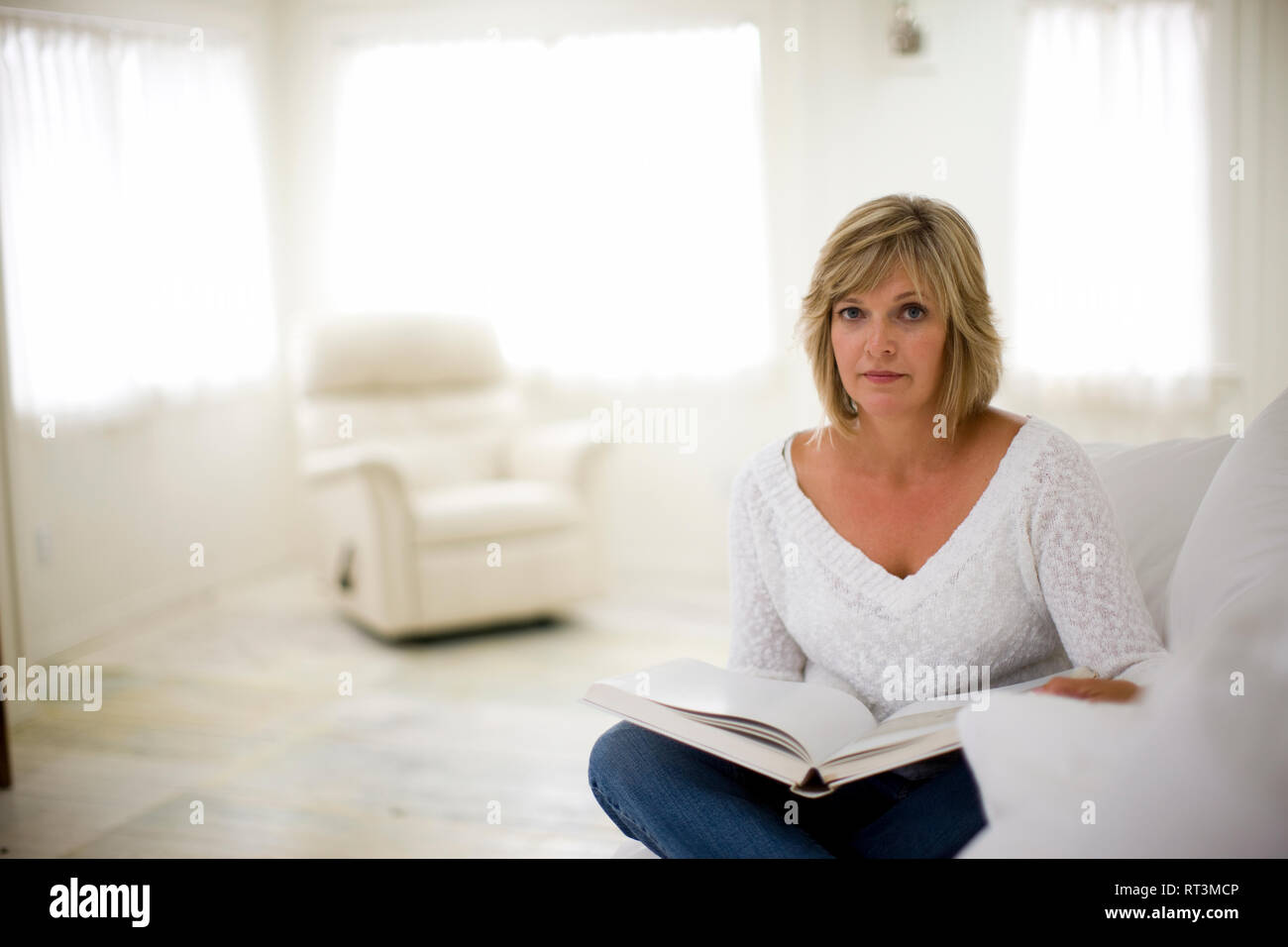 Portrait of a mid-adult woman sitting with a book on a couch inside her home. Stock Photo