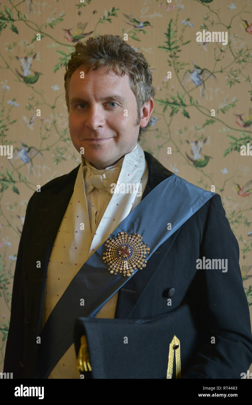 A handsome man dressed as a Victorian office wearing expensive clothes and a Royal sash stands for a. portrait. He looks serious and thoughtful Stock Photo