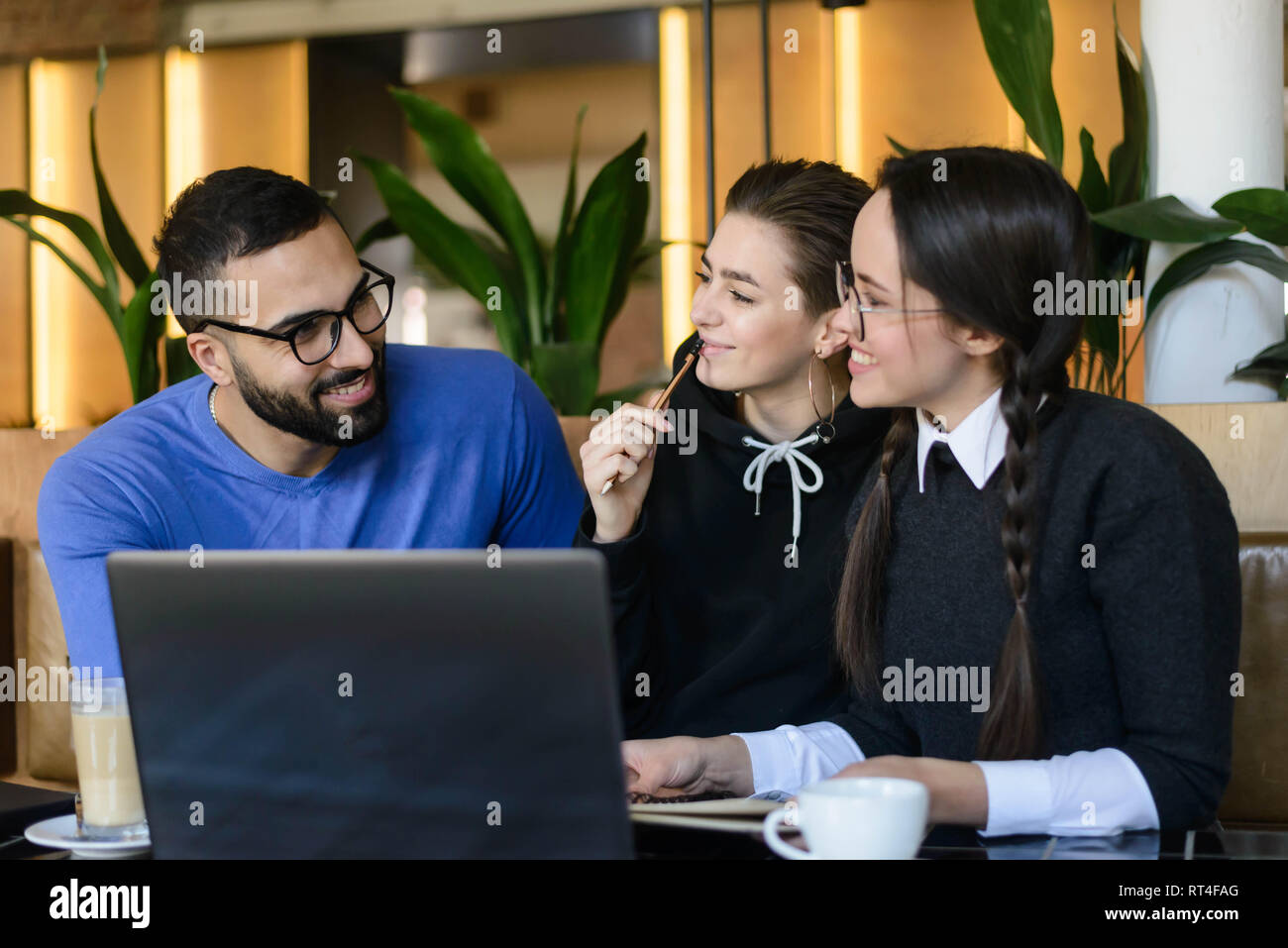 Group of joyful positive students talking, enjoying and using laptop in cafe Stock Photo