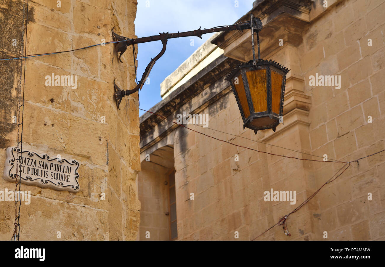 Panoramic view on a old, antic, medieval and historic lantern hanging on a sand stone wall. In the Background the castle of Mdina, Malta. Europe Stock Photo