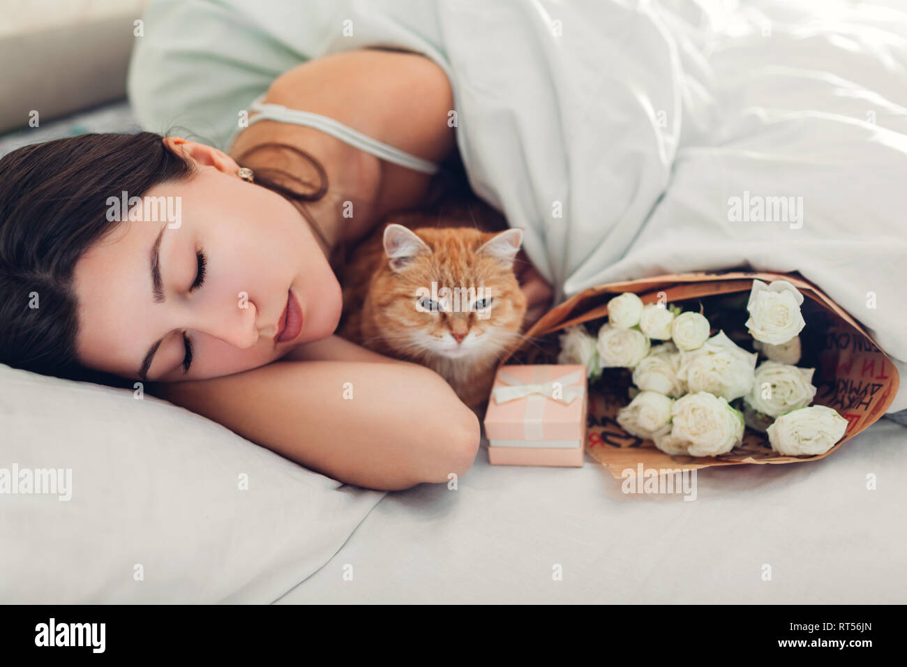 Young woman sleeping near bouquet of roses and gift box in bed with cat. Present left by boyfriend for Women's day. Surprise Stock Photo