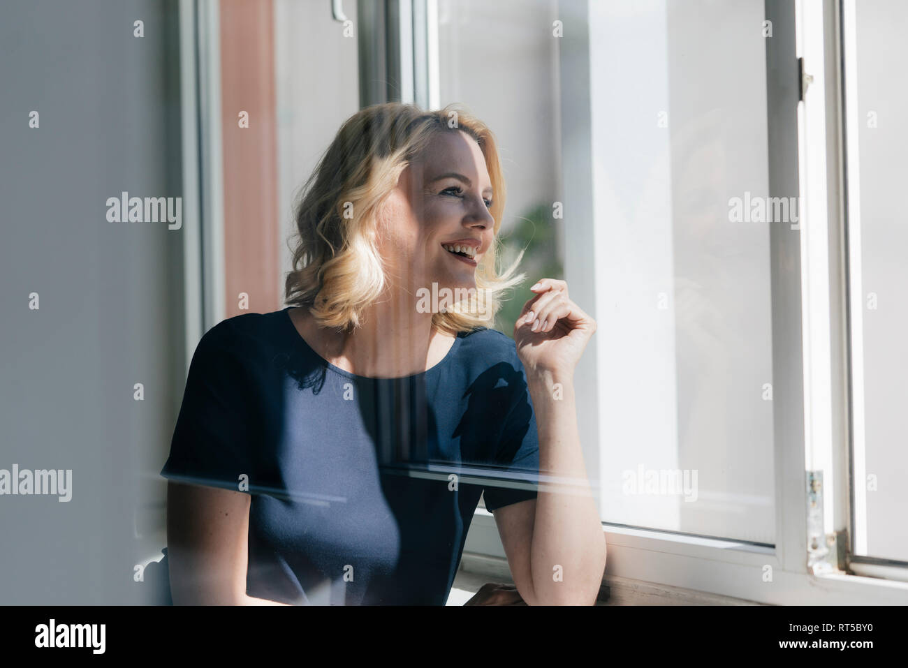 Happy blond woman in sunlight looking out of window Stock Photo - Alamy