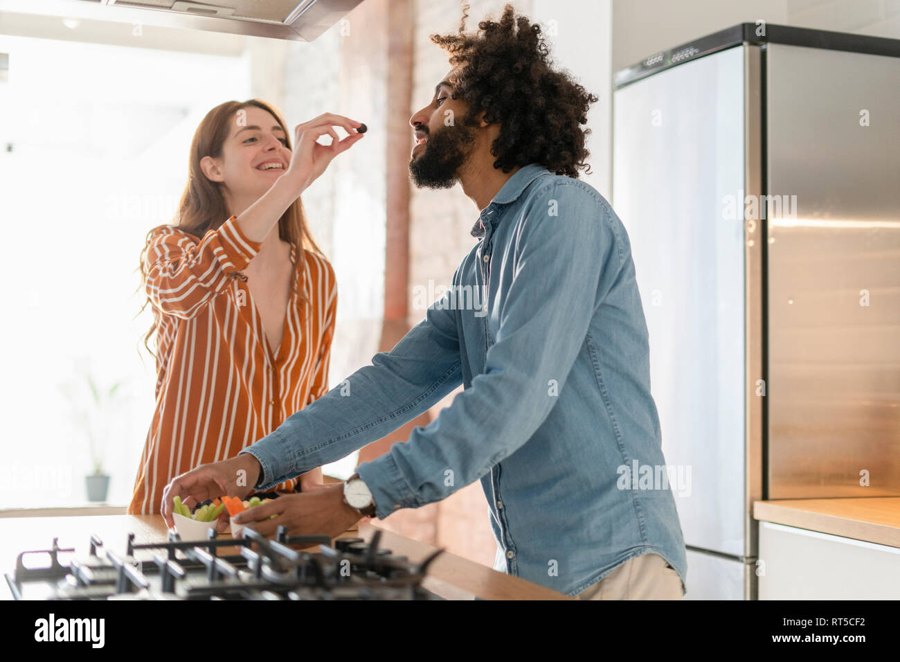 Couple standing in kitchen, preparing dinner party, woman feeding man with olive Stock Photo