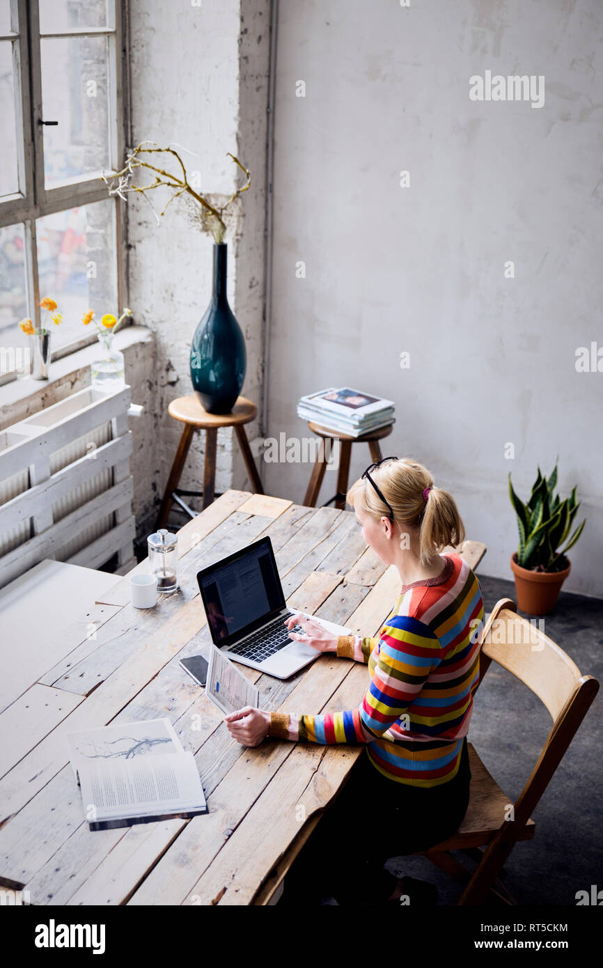 Woman sitting at desk in a loft using laptop and tablet Stock Photo