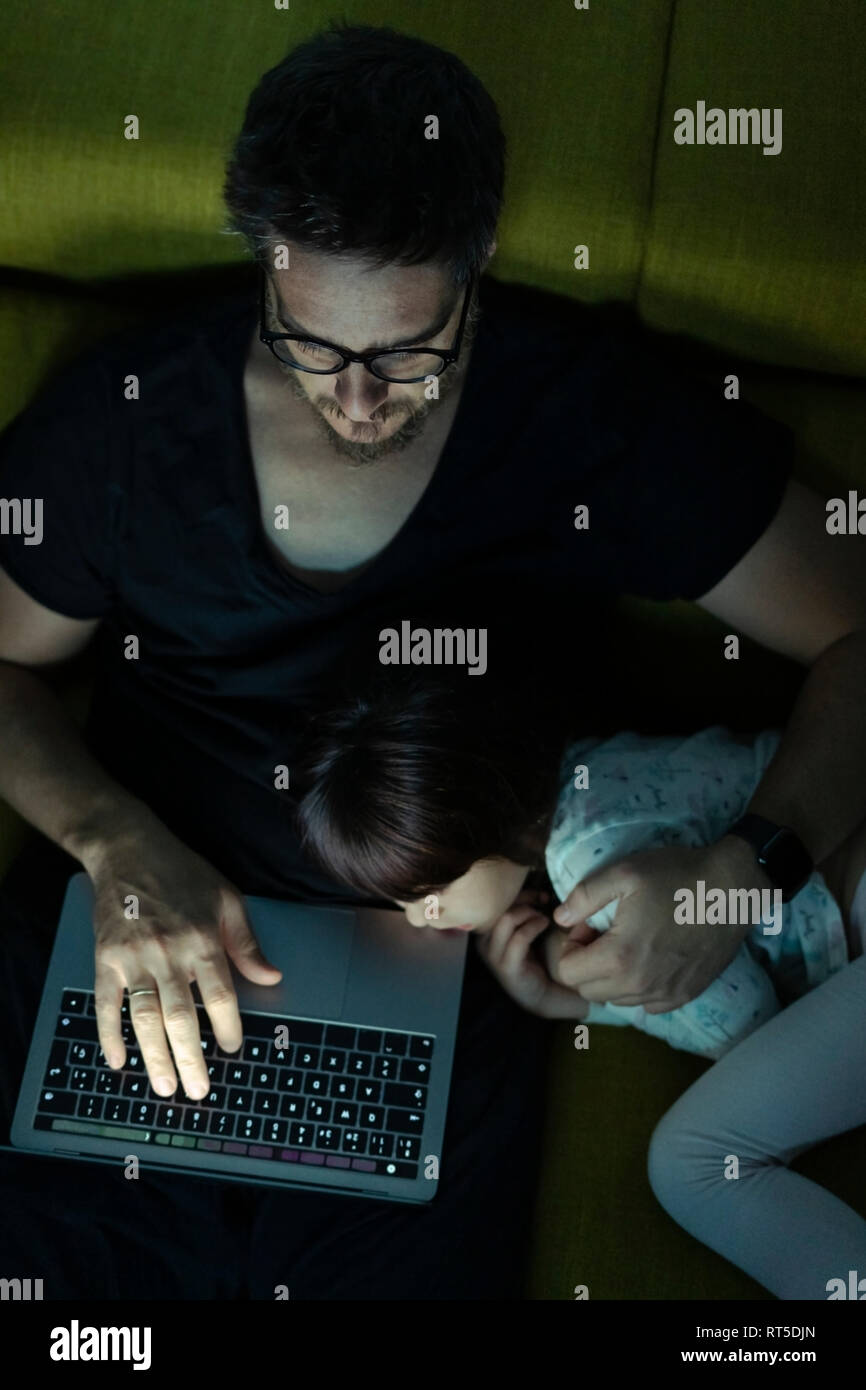 Father using laptop on couch at night with daughter  lying next to him Stock Photo