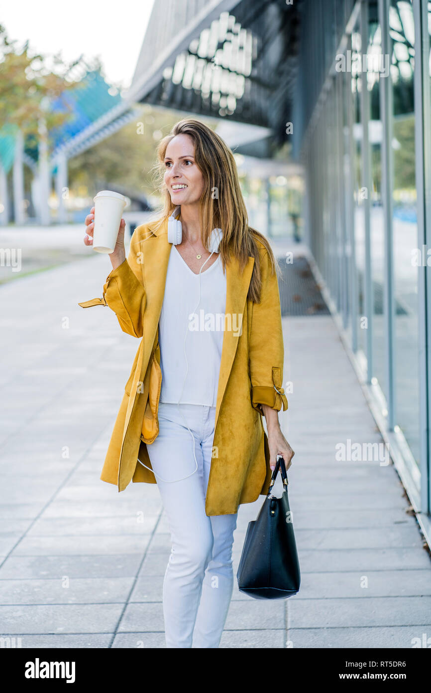 Smiling woman with handbag and takeaway coffee walking along building Stock Photo