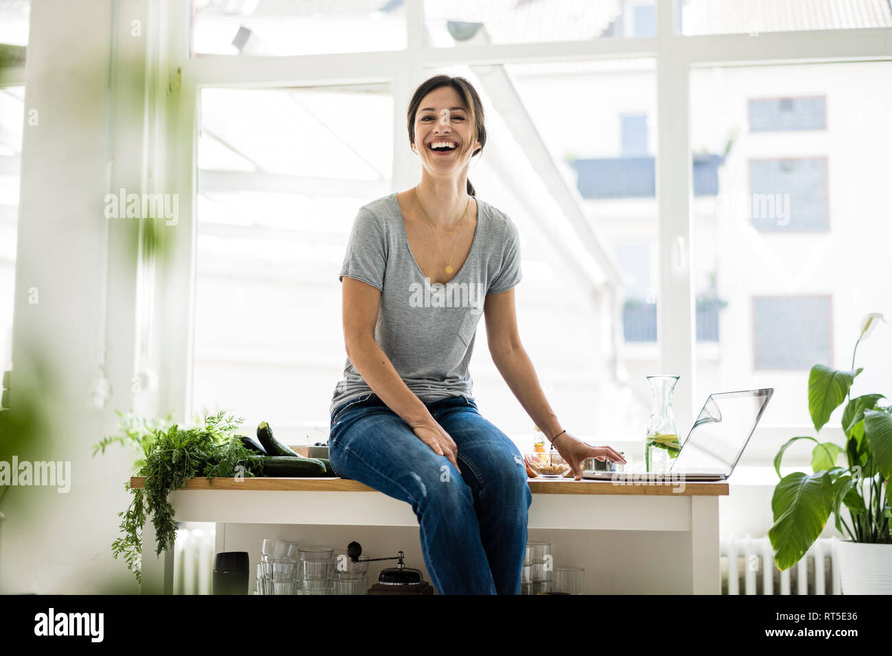 Woman sitting on kitchen table, searching for healthy recipes, using laptop Stock Photo