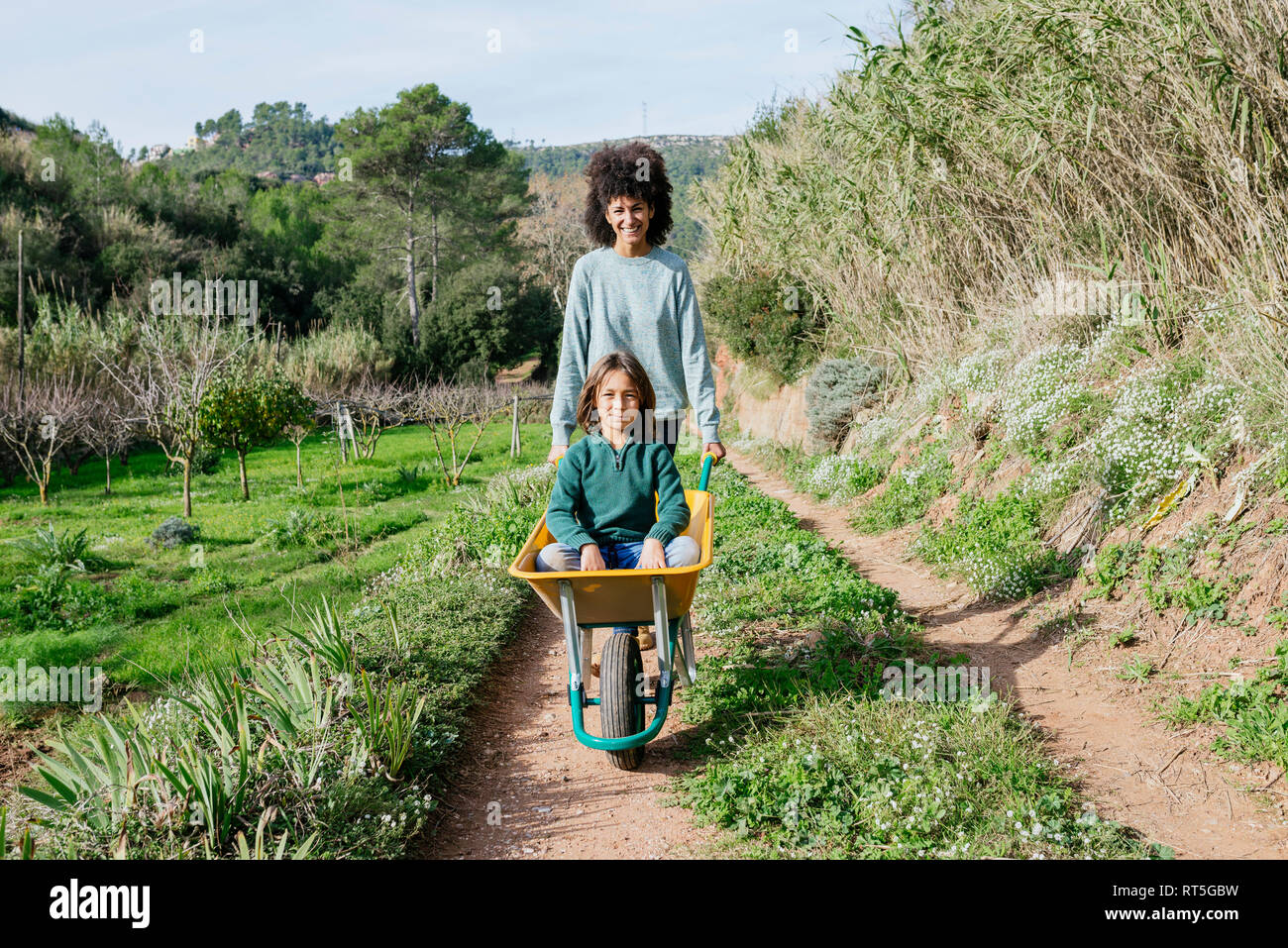 Mother walking on a dirt track, pushing wheelbarrow, with his son sitting in it Stock Photo