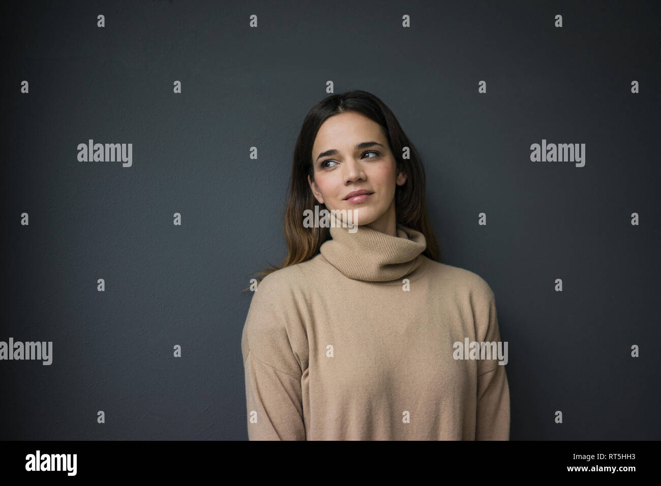 Portrait of smiling woman wearing light brown turtleneck pullover leaning against grey wall Stock Photo