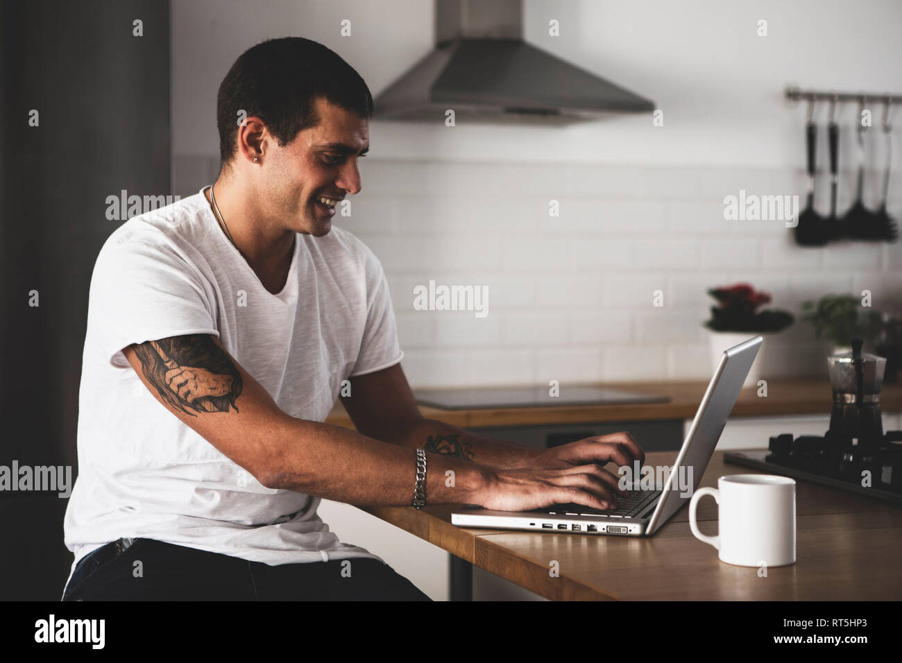 Smiling young man using laptop in kitchen at home Stock Photo