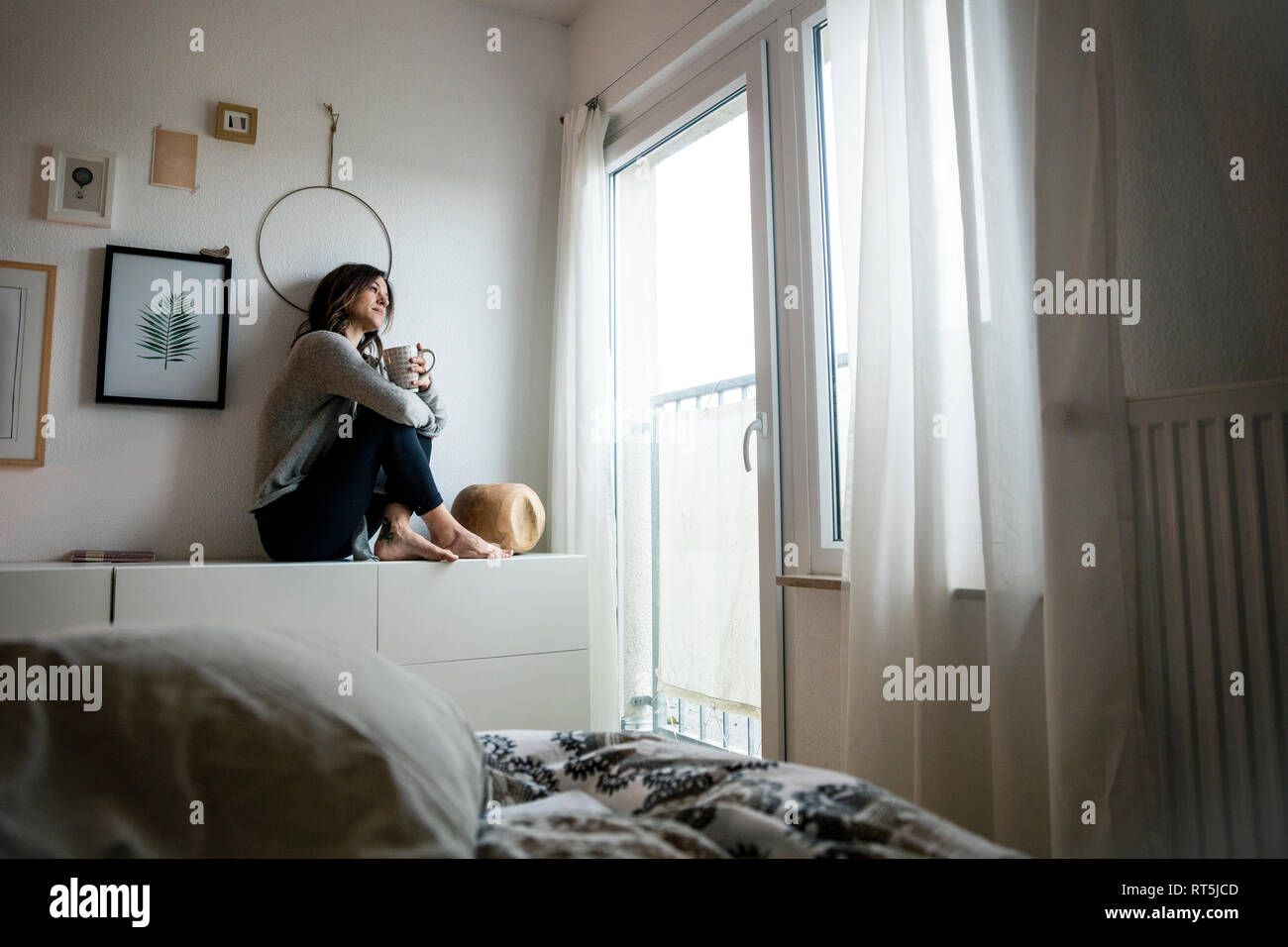Woman sitting on chest of drawers, drinking tea, relaxing Stock Photo