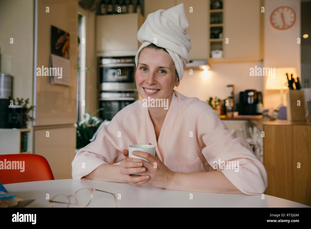Portrait of smiling woman wearing a towel turban sitting with cup of coffee at table in the kitchen Stock Photo