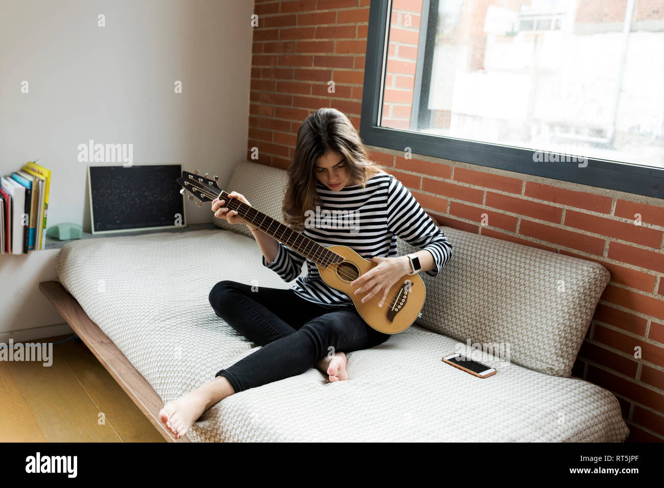 Young woman sitting on couch at home playing guitar Stock Photo