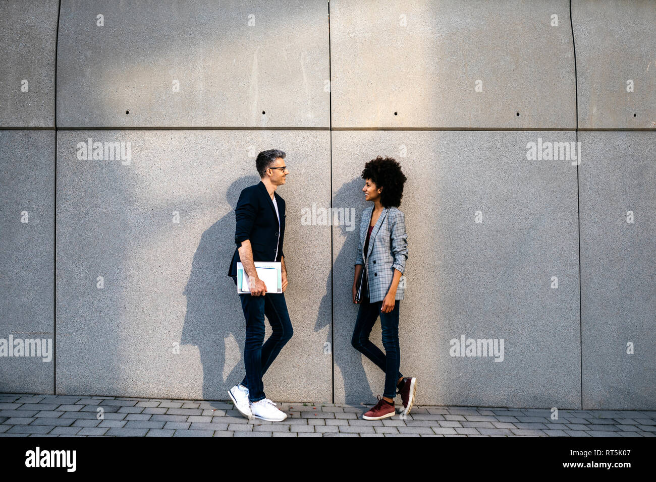 Two colleagues talking outdoors leaning against a wall Stock Photo