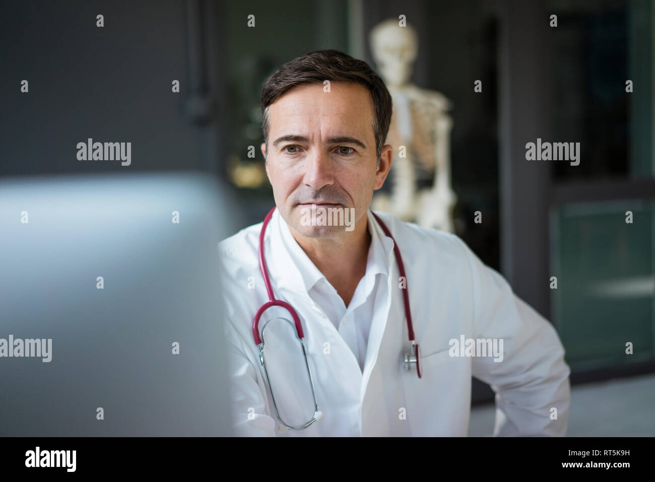 Doctor in medical practice looking at computer with skeleton in background Stock Photo