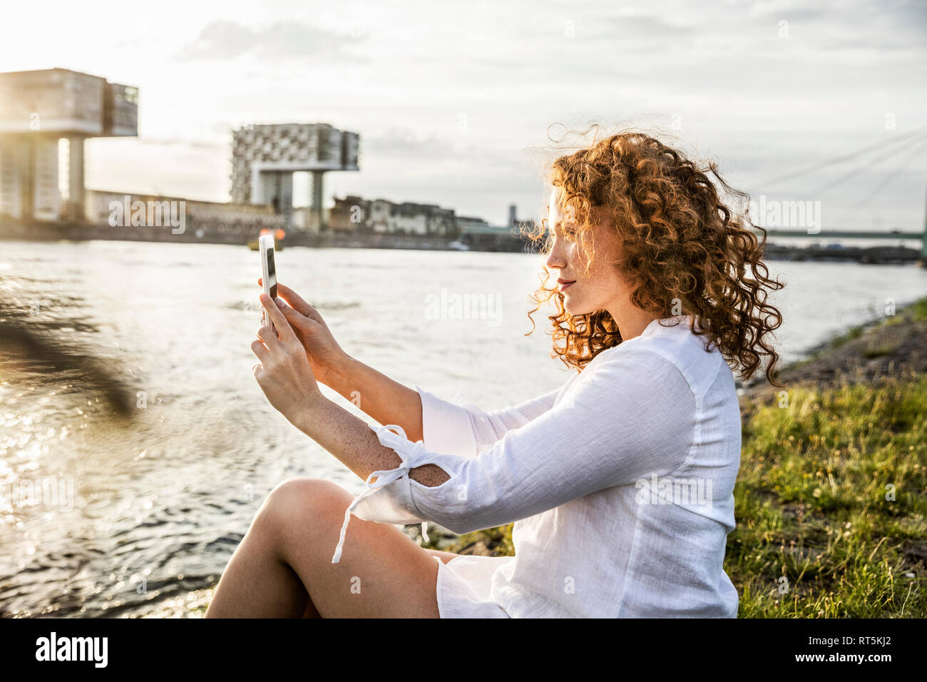 Germany, Cologne, young woman sitting at riverside in the evening taking selfie with cell phone Stock Photo