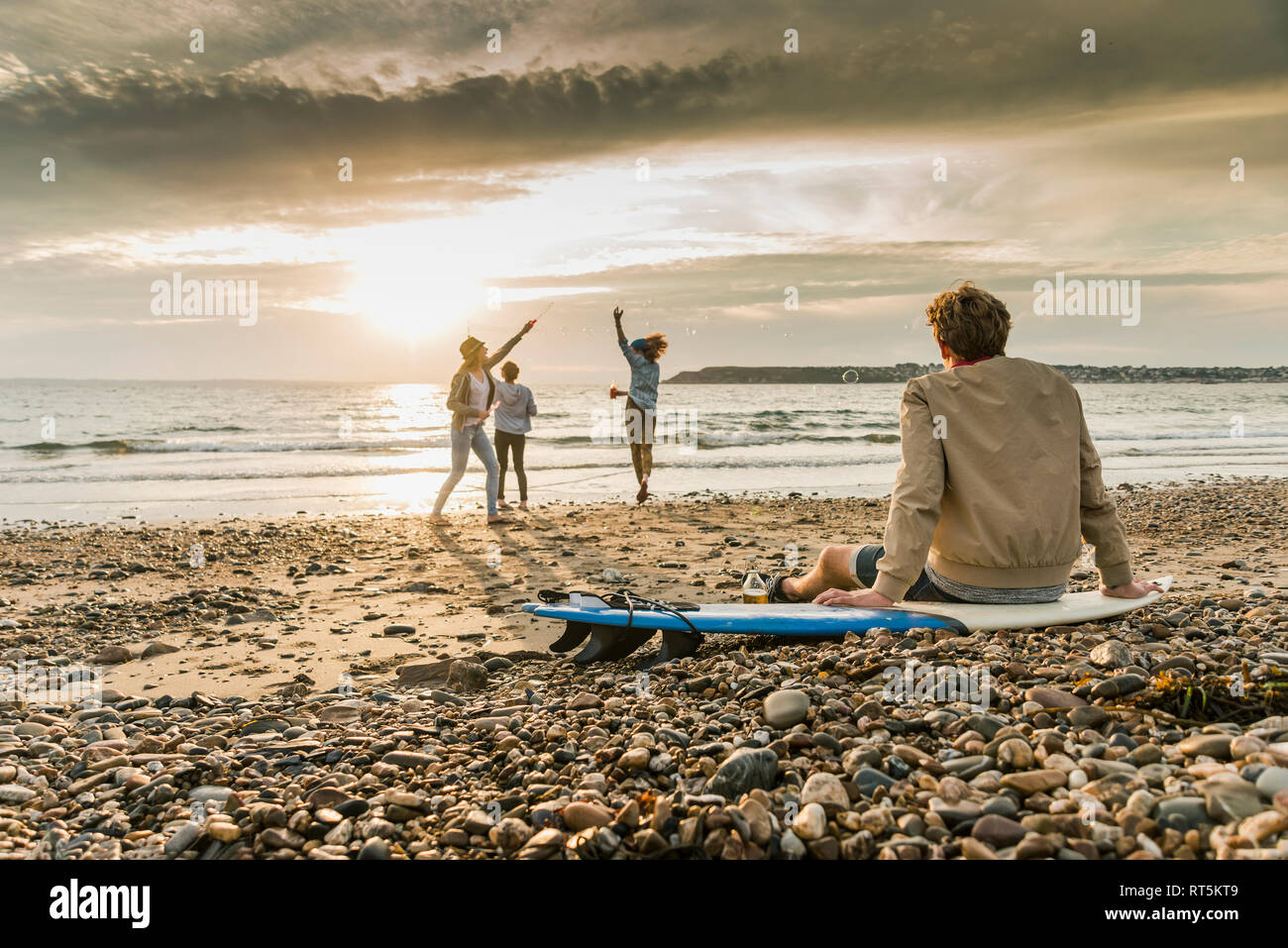 Young man on surfboard watching friends making soap bubbles on the  beach at sunset Stock Photo
